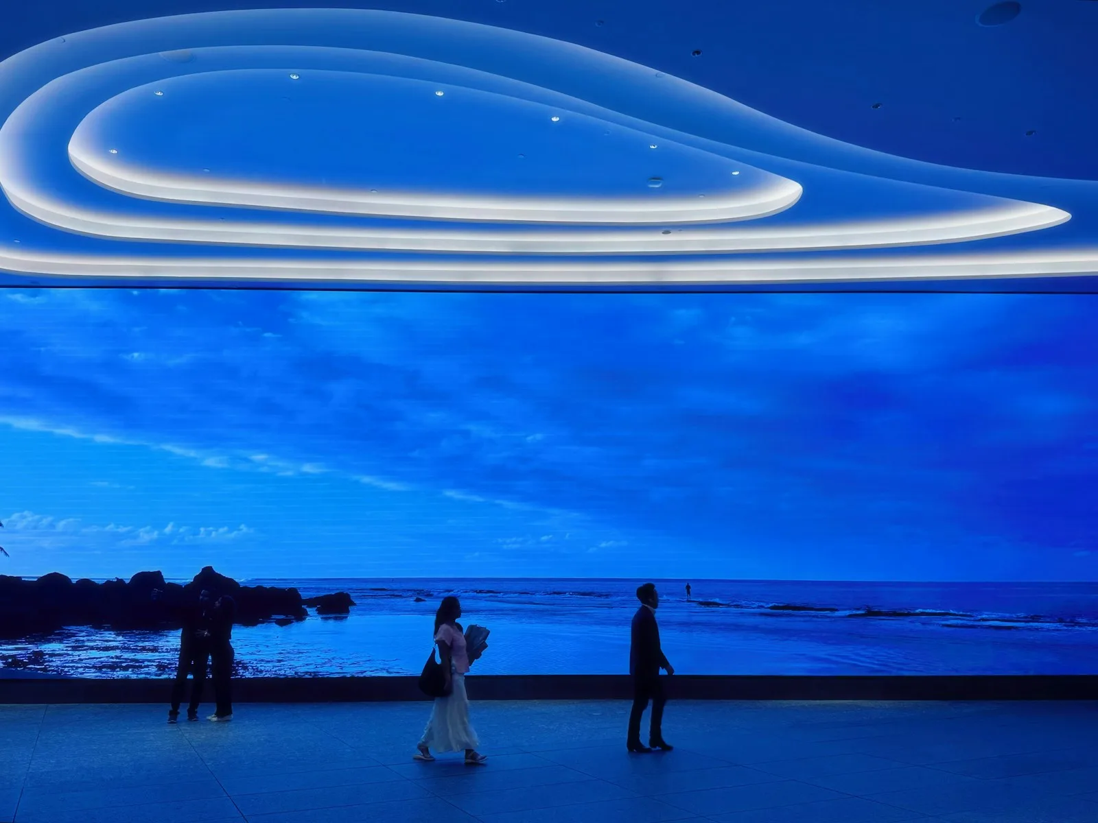 곡선 조명 천장 아래 바다 풍경을 띄운 대형 LED 벽 앞을 지나가는 사람들